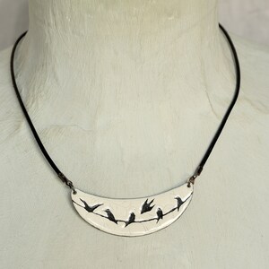 May include: A white ceramic necklace with a crescent moon pendant featuring a black silhouette of birds perched on a branch.