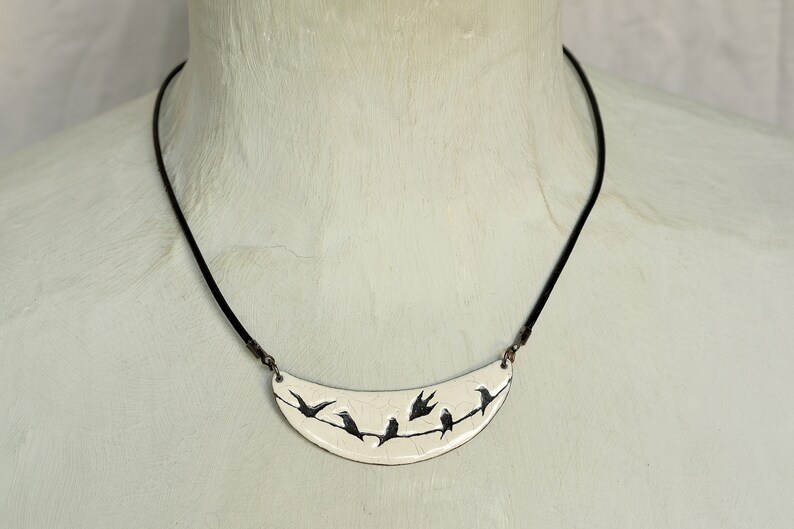 May include: A white ceramic necklace with a crescent moon pendant featuring a black silhouette of birds perched on a branch.