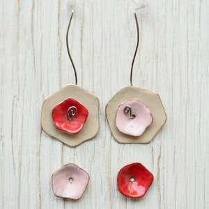 Peut inclure: Une paire de boucles d'oreilles pendantes avec une base en céramique blanche et un motif de fleur rose et rouge. Il y a aussi deux boucles d'oreilles plus petites avec le même motif de fleur.