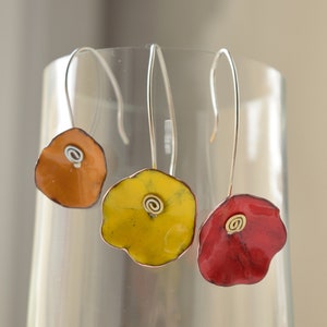 Peut inclure: Trois paires de boucles d'oreilles avec des pendentifs en forme de fleur. Les pendentifs sont en métal et ont une surface texturée. Les boucles d'oreilles sont disponibles en orange, jaune et rouge.