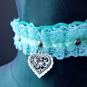 May include: A light blue lace choker with a silver heart charm. The heart charm has a decorative design with a butterfly.