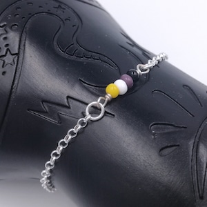 Op de afbeelding: Een zilveren kettingarmband met vier kleine kralen in de kleuren geel, wit, paars en zwart.