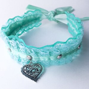 May include: A light blue lace choker with a silver heart charm. The choker has a satin ribbon tie and silver accents.