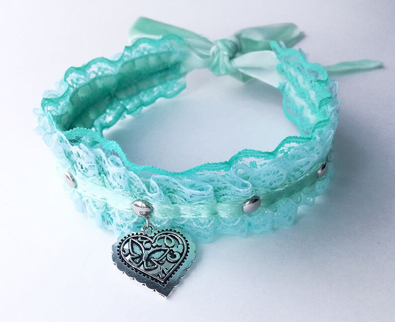 May include: A light blue lace choker with a silver heart charm. The choker has a satin ribbon tie and silver accents.