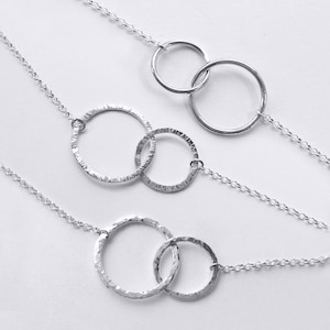 May include: Three silver necklaces, each featuring two interlocking circular pendants. One necklace has smooth, polished rings, while the other two necklaces display rings with a textured, hammered finish. All pendants are attached to delicate silver chains.