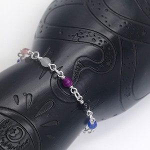 Op de afbeelding: Een zilveren kettingarmband met vijf gekleurde edelsteen kralen. De kralen zijn roze, lichtblauw, paars, zwart en blauw.