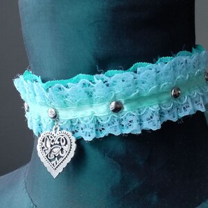 May include: A light blue and green lace choker with a silver heart charm. The choker has silver studs along the ribbon.