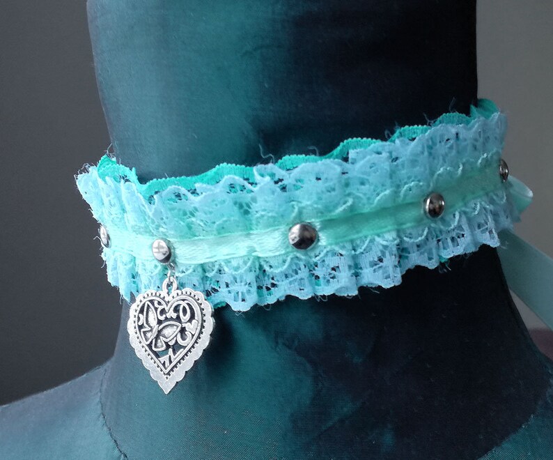 May include: A light blue and green lace choker with a silver heart charm. The choker has silver studs along the ribbon.