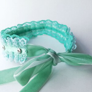 May include: A light green lace choker with a matching satin ribbon tied in a bow. The choker has two silver metal accents.