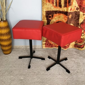 Könnte beinhalten: Zwei rote gepolsterte Hocker mit schwarzen Metallfüßen. Die Hocker haben eine quadratische Oberseite und ein schlichtes, modernes Design.