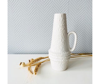 May include: White ceramic vase with a textured surface and a handle. The vase has a narrow opening and a wide base.