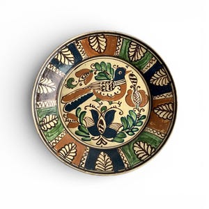 May include: A round, hand-painted ceramic plate with a bird and floral design. The plate features a cream-colored center with a brown bird, blue accents, and green leaves. The border has alternating sections of brown, green, and blue with leaf patterns.
