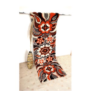 Peut inclure: Un tapis floral vintage avec un motif répété de grandes fleurs orange, blanches et marron. Le tapis est suspendu à une barre métallique. Un vase décoratif avec de l'herbe séchée est à gauche.