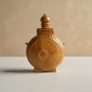 May include: A brown ceramic flask with a decorative geometric pattern. The flask has a round body, a small handle on each side, and a stopper on top. The base has two small feet.