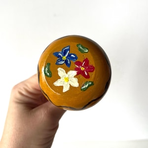 May include: A wooden knob with a brown base and a black outline. The knob is painted with three flowers: a blue flower, a red flower, and a white flower.
