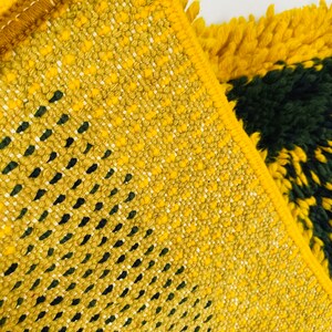 Op de afbeelding: Close-up van een geweven tapijt in geel en groen met een geometrisch patroon. Het tapijt heeft een franje rand.