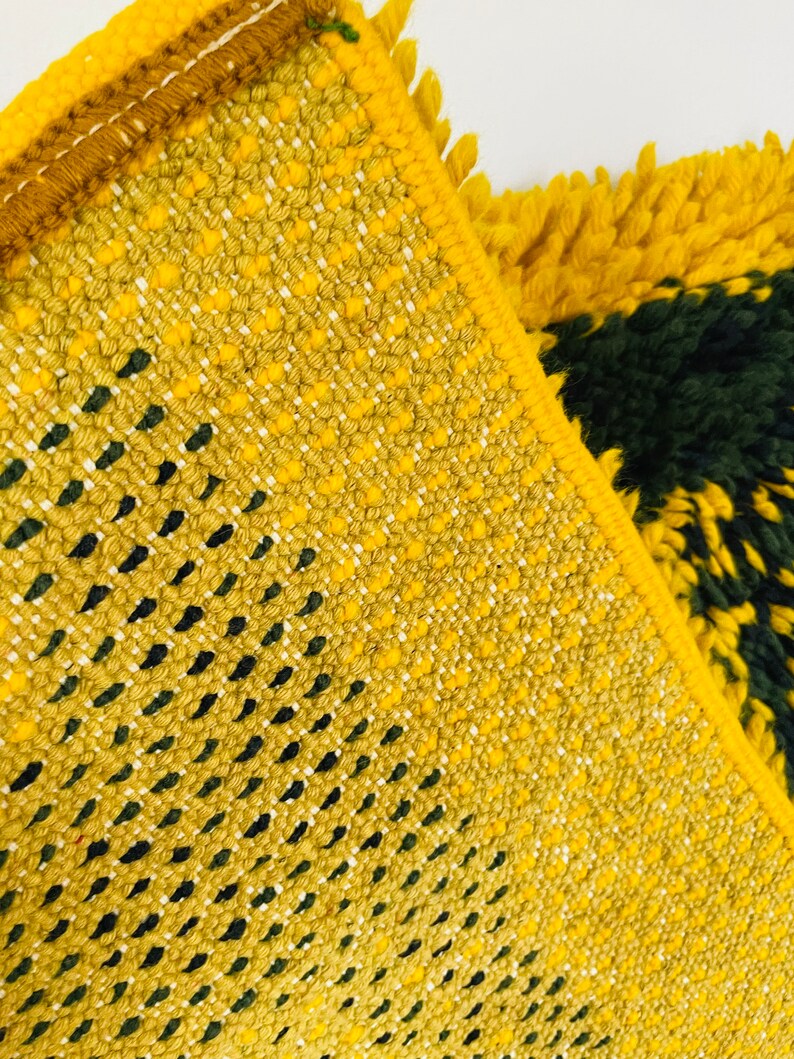 Op de afbeelding: Close-up van een geweven tapijt in geel en groen met een geometrisch patroon. Het tapijt heeft een franje rand.