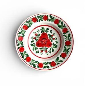 May include: A white ceramic plate with a hand-painted floral design. The plate features a red and green colour scheme, with red berries and green leaves arranged in a circular pattern around the rim and centre.