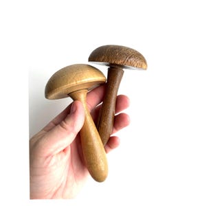 May include: Two wooden mushroom-shaped objects. One is light-colored, the other is dark brown. The mushroom caps are rounded, and the stems are smooth. The objects are held in a hand against a white background.