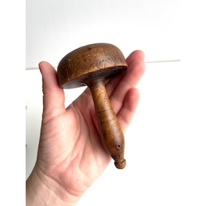 May include: A vintage wooden mushroom-shaped tool with a rounded top and a long, slender handle. The wood has a warm, brown tone with visible grain and a small hole near the handle's end. The tool is held in a hand against a white background.