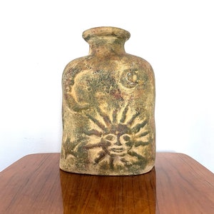 May include: A ceramic vase with a rustic brown and green glaze. The vase features a sun and moon design with a face on the sun.