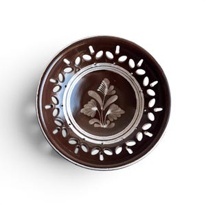 Può includere: Un piatto in ceramica marrone con un motivo floreale al centro. Il piatto ha un bordo bianco e motivi decorativi ritagliati sul bordo. Il motivo floreale è in una tonalità più chiara di marrone.