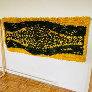 Op de afbeelding: Een geel en groen shag-kleed met een geometrisch patroon. Het kleed hangt aan een witte muur.