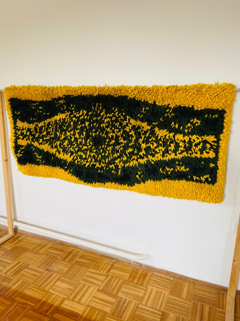 Op de afbeelding: Een geel en groen shag-kleed met een geometrisch patroon. Het kleed hangt aan een witte muur.