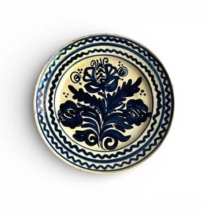 Puede incluir: Plato de cerámica redondo con un centro color crema y un diseño floral azul oscuro. El borde presenta un patrón azul ondulado. El diseño del plato incluye flores y hojas estilizadas, creando una pieza decorativa y artística.