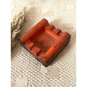 Op de afbeelding: Een vierkante keramische asbak met een levendig oranje glazuur en een textuurrijke, gespikkelde afwerking. Het object heeft afgeronde randen en vier duidelijke inkepingen aan de zijkanten om items op te laten rusten, met een diepe centrale kom.