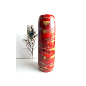 Op de afbeelding: Een hoge, cilindervormige keramische vaas met een glanzende afwerking. De vaas heeft een levendig kleurenpalet van rood, geel en bruin, met een marmer effect. Een pauwenveer staat naast de vaas.