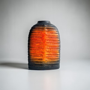 Op de afbeelding: Een keramische vaas met een textuur, geribbeld ontwerp. De vaas heeft een kleurovergang, die overgaat van een donkerzwarte basis naar levendige oranje en rode tinten in het midden. De bovenkant is zwart.