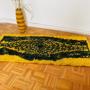 Op de afbeelding: Een geel en groen shag-kleed met een geometrisch patroon. Het kleed is gemaakt van dikke, pluche vezels en heeft een modern ontwerp.