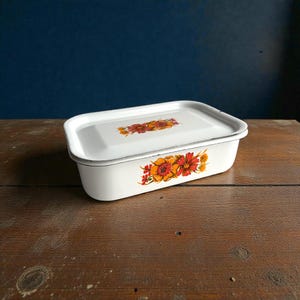 Puede incluir: Contenedor rectangular blanco de esmalte con tapa. El contenedor presenta un diseño floral en tonos naranja, rojo y amarillo. El contenedor está sobre una superficie de madera.