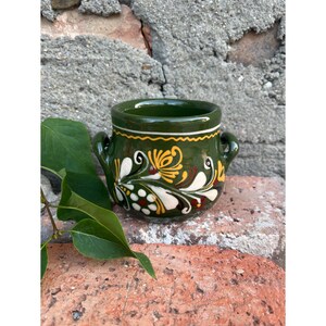 Può includere: Una piccola tazza in ceramica fatta a mano con base verde scuro e due piccole maniglie. La tazza è adornata da un motivo floreale in bianco, giallo e marrone. Una linea ondulata bianca e gialla decora il bordo.