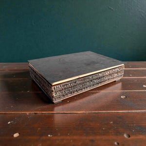 Può includere: Una scatola di legno scuro rettangolare con un design strutturato e stratificato sui lati. La scatola è su una superficie di legno, con uno sfondo verde scuro. La scatola sembra essere vintage o antica.