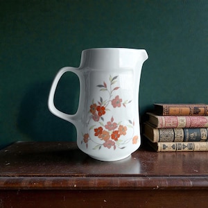 Puede incluir: Jarra de cerámica blanca con asa curva y pico vertedor. La jarra está decorada con un estampado floral en tonos naranja y rosa. La jarra está sobre una superficie de madera. Altura aproximada: 20 cm.