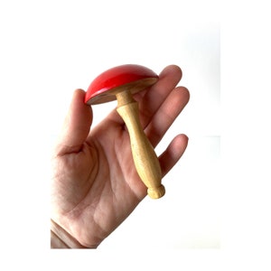 May include: A red and wooden toy mushroom with a stem and a rounded top. The mushroom is held in a hand.