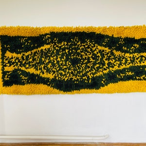 Op de afbeelding: Een geel en groen shag-kleed met een geometrisch patroon. Het kleed hangt aan een witte muur.