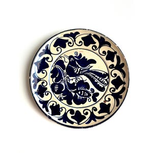 May include: Round ceramic plate with a hand-painted design. The plate features a central image of a bird perched on a branch, surrounded by blue floral and scrollwork patterns against a cream background. The rim is also decorated with blue designs.