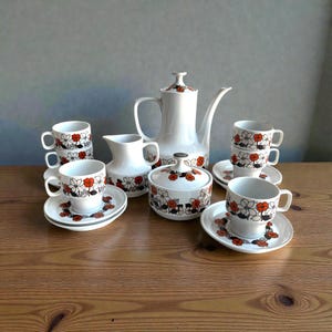 May include: A vintage white ceramic tea set with a floral pattern in orange, black, and white. The set includes a coffee pot, creamer, sugar bowl, cups, and saucers, arranged on a wooden surface.