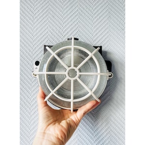 May include: A circular industrial light fitting with a clear, ribbed glass lens and a white, radial grid. The fitting is mounted on a black metal base. The light is held by a hand.