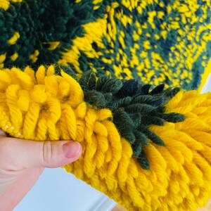 Op de afbeelding: Close-up van een geweven tapijt in geel en groen met een pluche textuur. Het tapijt is gemaakt van dik garen en heeft een uniek, abstract patroon.