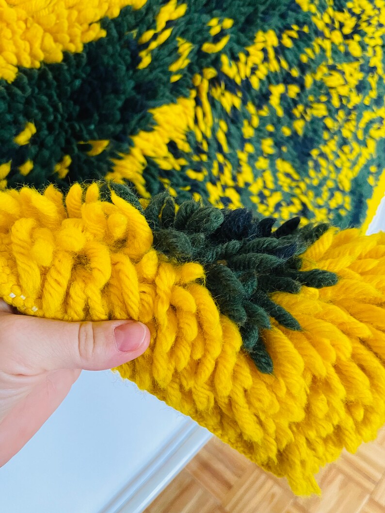 Op de afbeelding: Close-up van een geweven tapijt in geel en groen met een pluche textuur. Het tapijt is gemaakt van dik garen en heeft een uniek, abstract patroon.
