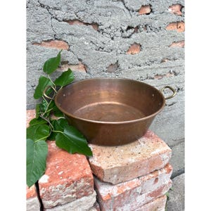 May include: A copper bowl with two small handles. The bowl is round and shallow. It rests on a stack of red bricks, with green leaves and a textured grey wall in the background. The bowl's diameter is approximately 30 cm.