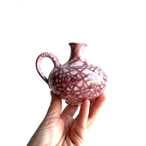 May include: A small, round, ceramic jug with a handle and a narrow neck. The jug is white with a cracked, web-like pattern in shades of burgundy. The jug is held in a person's hand.