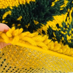 Op de afbeelding: Close-up van een geweven tapijt in geel en groen met een pluche textuur. Het tapijt heeft een geometrisch patroon en is gemaakt van dik garen.