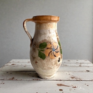 May include: A vintage ceramic pitcher with a handle, featuring a cream-colored base with brown and tan accents. The pitcher is adorned with a hand-painted floral design in orange, green, and blue. The rim and handle have a brown glaze.