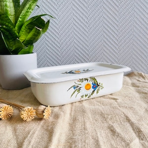 May include: White enamelware baking dish with a floral design featuring white daisies, blue flowers, and green leaves. The dish has a lid.