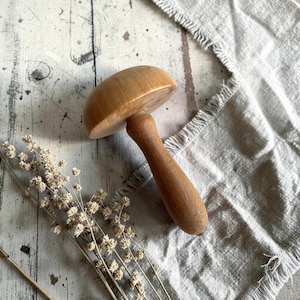 May include: A wooden mushroom-shaped object with a rounded top and a handle. The mushroom is light brown, and the handle is a darker shade of brown. The object is placed on a distressed, light-colored wooden surface, with dried flowers and a linen cloth.